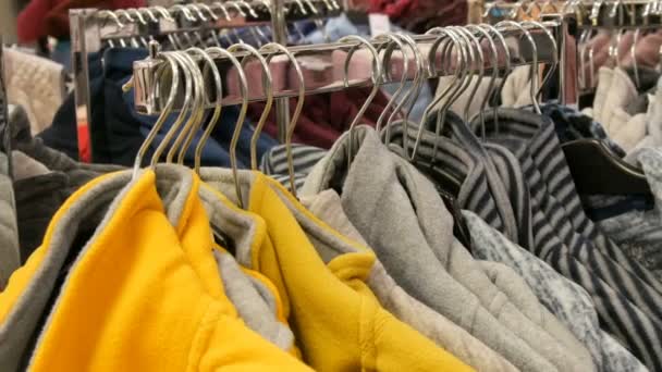 Gran número de suéteres de invierno cálidos multicolores cuelgan de las perchas en la tienda del centro comercial, los compradores pasan
. - Imágenes, Vídeo