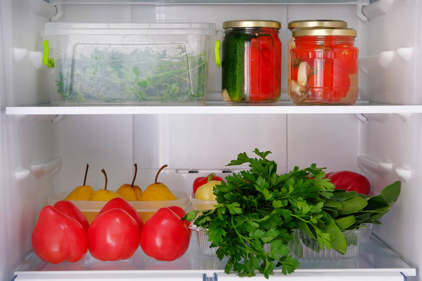 Open koelkast. Gezonde rauwkost: peren, komkommers, tomaten, peterselie, peper. Conserven in kruik en groenten op plank van koelkast. - Foto, afbeelding
