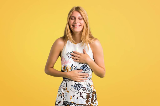 Blonde young girl in a summer dress smiling a lot while putting hands on chest on yellow background - Fotografie, Obrázek