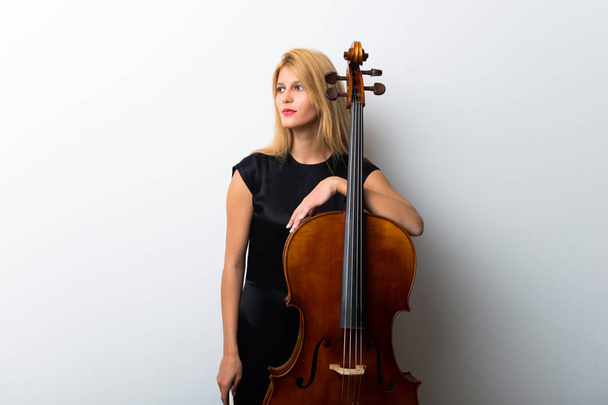 Young blonde girl with her cello posing on white wall - 写真・画像