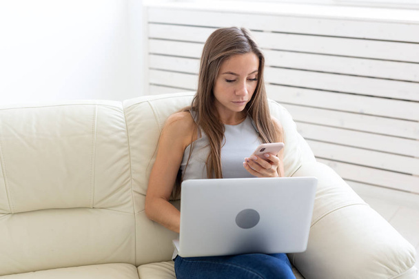 Concepto de tecnologías, ocio y personas: mujer joven sentada en el sofá blanco y charlando en el netbook y en el móvil
 - Foto, Imagen
