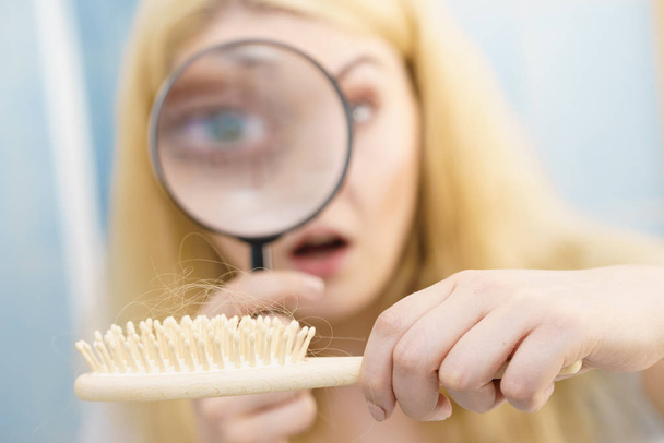 Woman using magnifying glass looking at her brush. Examing hair loss, haircare concept. - 写真・画像