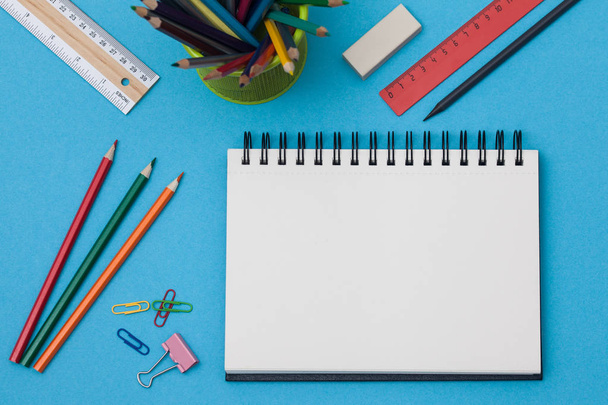 Empty spiral notebook with white page and multicolored stationery on a blue table. Workplace with notepad on colored background. Sketch-book for input the text, top view. Back to school concept. - Fotó, kép