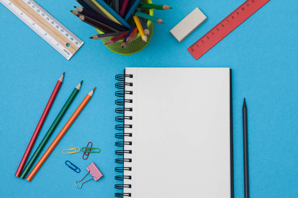 Empty spiral notebook with white page and multicolored stationery on a blue table. Workplace with notepad on colored background. Sketch-book for input the text, top view. Back to school concept. - Foto, imagen