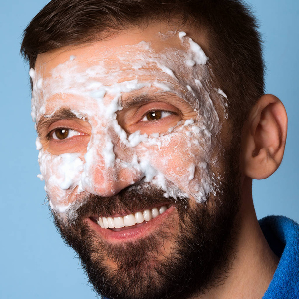 der lächelnde Mann mittleren Alters benutzt Schaum zur Hautpflege auf seinem Gesicht auf blauem Hintergrund. - Foto, Bild