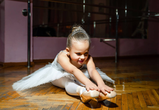 Attractive young ballerina wearing white tutu stretching to her pointe shoes sitting on the wooden floor. Little girl doing exercises in studio - Photo, image