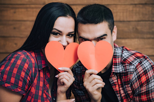Fun being together. Young lovely couple sitting on the bed and hiding their faces with red paper hearts over wooden wall background. Close-up - Zdjęcie, obraz
