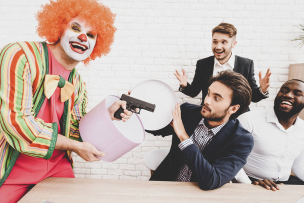 Young Man in Clown Costume on Meeting in Office. April Fools Day. Man with Gift Box. April Jokes. Toy Gun. Workers on Meeting. Holidays and Celebration Concept. Clown with Red Nose. - 写真・画像