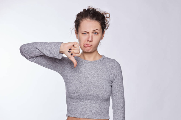 Discontent attractive European woman shows disapproval sign, keeps thumb down, expresses dislike, frowns face in discontent, dressed in gray top, isolated over white background. Body language concept. - Fotografie, Obrázek