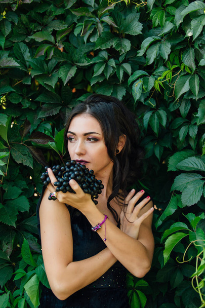 Portrait of young sexy brunette in black dress with black grapes. - Photo, Image