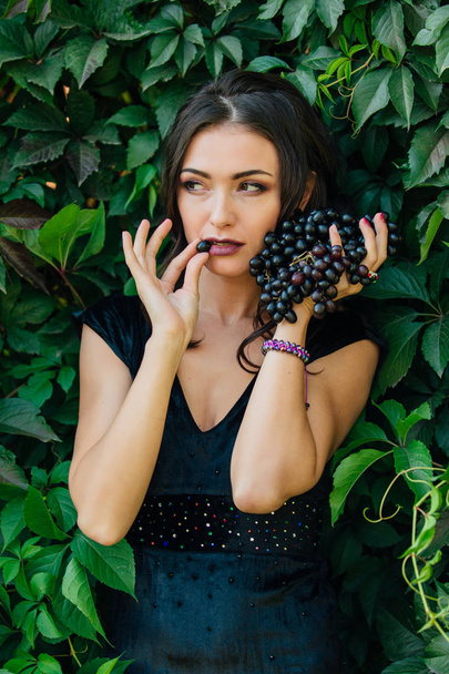 Portrait of young sexy brunette in black dress biting black grapes. - Фото, изображение