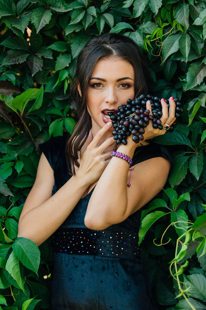 Portrait of young sexy brunette in black dress biting black grapes. - Photo, image
