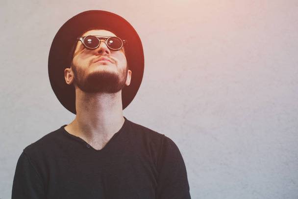 Porträt eines Hipster-Typen, der über weißen Hintergrund nach oben blickt. mit Sonnenbrille und schwarzem Hut - Foto, Bild