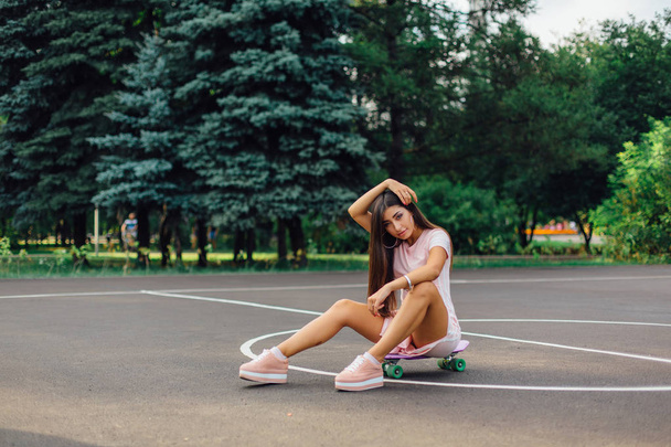 Portrait of a smiling charming brunette female sitting on her skateboard on a basketball court. Happy woman with trendy look taking break during sunset. - 写真・画像