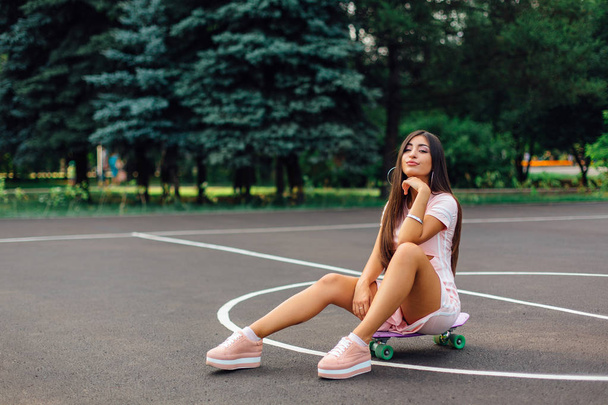 Portrait of a smiling charming brunette female sitting on her skateboard on a basketball court. Happy woman with trendy look taking break during sunset. - Foto, afbeelding
