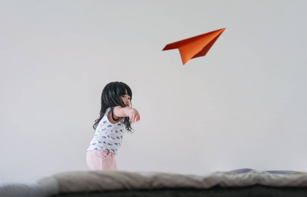 Reisen für Kinder oder Fantasie-Konzept. fröhliches und aufgeregtes dreijähriges Mädchen hebt die Hand und wirft einen Papierflieger in ein weißes Zimmer - Foto, Bild