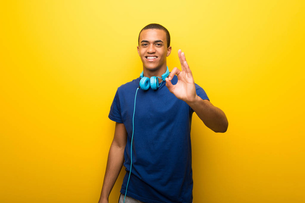 Hombre afroamericano con camiseta azul sobre fondo amarillo mostrando un signo aceptable con los dedos
 - Foto, Imagen