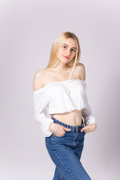 Studio shot of gorgeous young blonde woman with straight hair wearing off shoulder crop top sweater and high waisted denim shorts. Gray isolated background, copy space, close up. - Foto, imagen