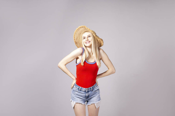 Studio shot of gorgeous young blonde woman with straight hair wearing wide brim straw hat, tight red tank top and sexy high waisted denim shorts. Gray isolated background, copy space, close up. - Zdjęcie, obraz