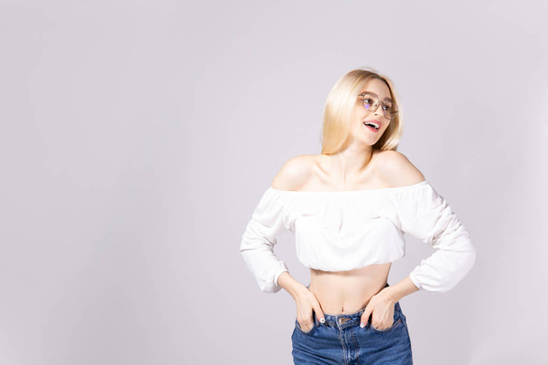 Studio shot of gorgeous young blonde woman with straight hair wearing off shoulder crop top sweater and high waisted denim shorts. Gray isolated background, copy space, close up. - Fotó, kép