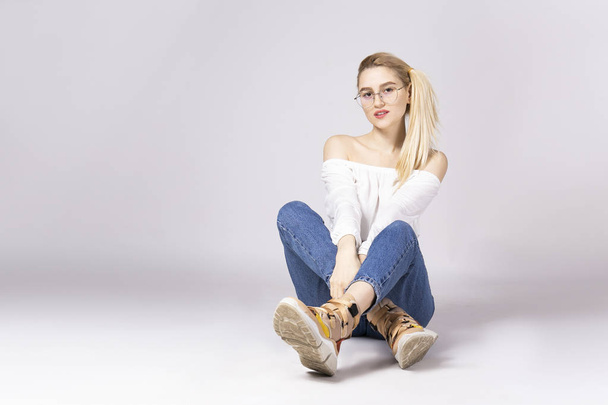 Studio shot of gorgeous young blonde woman with straight hair wearing off shoulder crop top sweater and high waisted denim shorts. Gray isolated background, copy space, close up. Ugly sneakers concept - Foto, Imagem