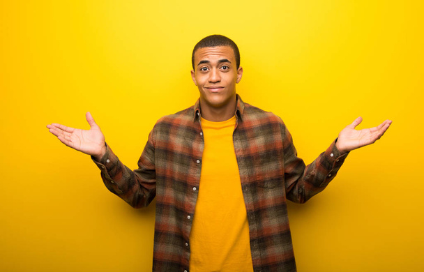Joven afroamericano sobre un vibrante fondo amarillo que tiene dudas mientras levanta las manos y los hombros
 - Foto, Imagen