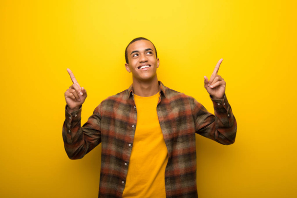 Νεαρός αφρικανική αμερικανική άνθρωπος σε ζωντανό κίτρινο φόντο, δείχνοντας με το δάχτυλο δεικτών μια μεγάλη ιδέα - Φωτογραφία, εικόνα