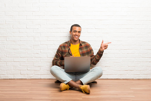 African american man sitting on the floor with his laptop pointing finger to the side in lateral position - 写真・画像