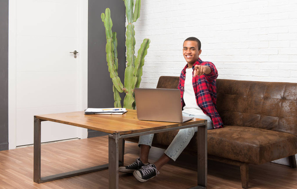 Hombre afroamericano con portátil en la sala de estar le señala con el dedo con una expresión de confianza
 - Foto, imagen