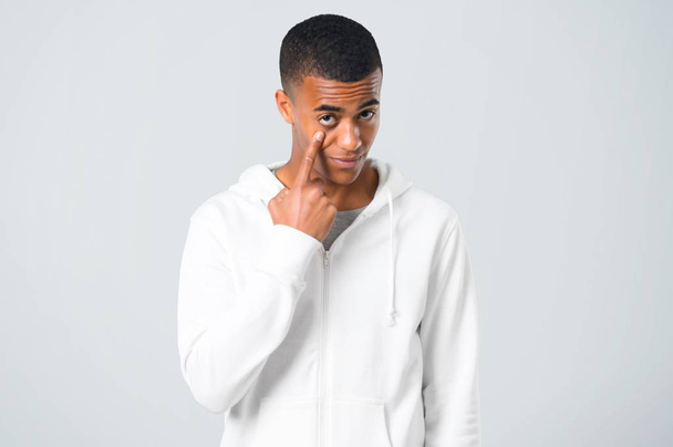 Μελαχροινός νεαρός με λευκό φούτερ στέκεται και κοιτάζοντας προς τα εμπρός, ανοίγοντας το μάτι με το δάχτυλο σε γκρίζο φόντο - Φωτογραφία, εικόνα