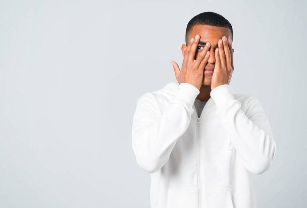 Dark-skinned young man with white sweatshirt covering eyes by hands and looking through the fingers on grey background - Фото, зображення