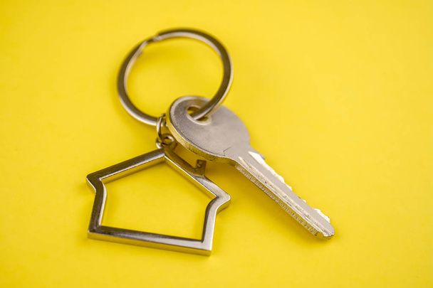das Konzept des Kaufs eines privaten Hauses oder einer Wohnung. Schlüsselanhänger aus Metall in Form eines Hauses mit einem Metallschlüssel auf gelbem Hintergrund. Nahaufnahme. - Foto, Bild
