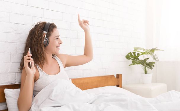 Szczęśliwa Kobieta w słuchawki do słuchania muzyki ze smartfona, gestykulacji w łóżku w domu, miejsce - Zdjęcie, obraz