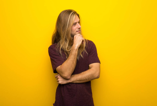 Blond man with long hair over yellow wall looking to the side with the hand on the chin - Foto, Bild
