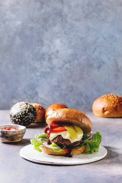 Variety of homemade classic and mini burgers in wheat and black buns with beef and veal cutlets, melted cheese and vegetables on white ceramic board over grey blue table. - Photo, Image