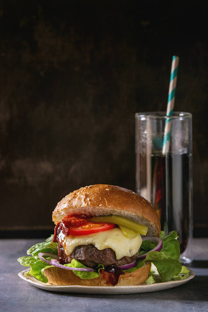 Close up of homemade classic burger in wheat bun with beef and veal cutlets, melted cheese and vegetables on white ceramic plate over dark table. Copy space. With glass of cola. - Фото, изображение