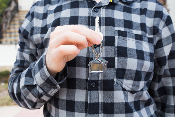Χέρι που κρατά το κλειδί με ένα μπρελόκ σε σχήμα του σπιτιού. Κλειδί σπίτι στο φόντο του νέου σπιτιού - Φωτογραφία, εικόνα
