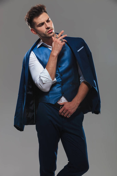 πορτρέτο του αλαζονική νεαρό άνδρα που φοράει το μπλε κοστούμι, κοιτάζοντας προς τα κάτω πλευρά ενώ στέκεται στο ανοιχτό γκρι φόντο - Φωτογραφία, εικόνα
