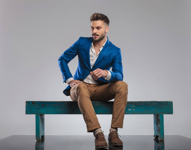 curious smart casual man sitting on wooden bench looks to side on grey background, full body picture - Foto, immagini