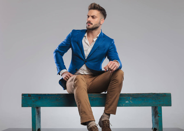 αλαζονική smart casual άνδρας κάθεται στο μπλε ξύλινο πάγκο φαίνεται πλευρά, πλήρη εικόνα σώματος - Φωτογραφία, εικόνα