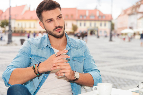 handsome casual man relaxing at table in city looks to side while sitting and fixing rings, portrait picture - Photo, Image