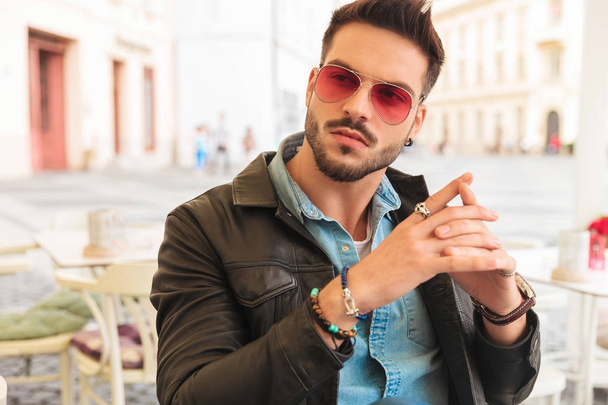 fashion man wearing sunglasses and holding palms together looks to side while sitting at table in city center - Foto, afbeelding
