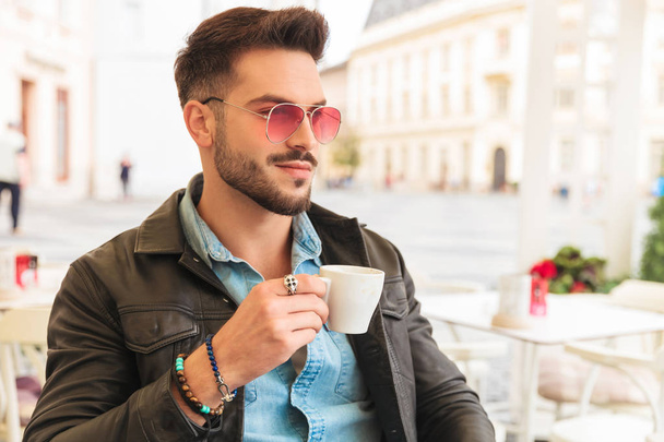 pohledný muž s přestávka na kávu na městské pozadí vypadá na stranu zatímco drží pohár a sedí u stolu, podobiznu - Fotografie, Obrázek