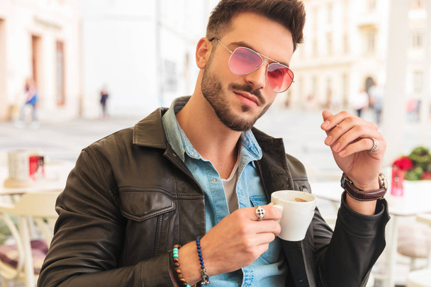 portrét atraktivní příležitostné člověka v kožené bundě pití kávy při sezení u stolu v městské pozadí - Fotografie, Obrázek