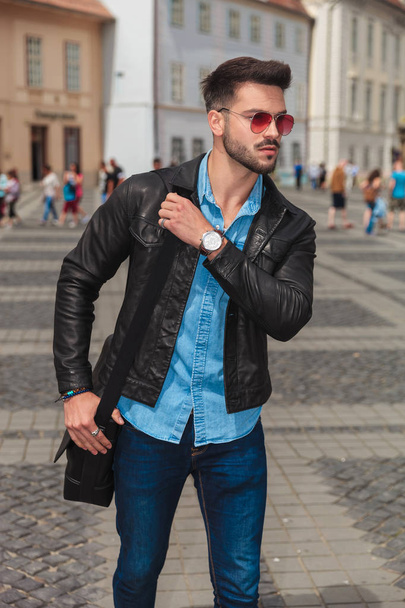 curious fashion man with red sunglasses going sightseeing the city and looking to side while holding shoulder bag - 写真・画像