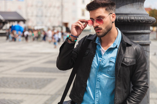 retrato de homem casual curioso fixando óculos de sol vermelhos enquanto estava na cidade com uma mão no bolso e encostado a um poste de iluminação
 - Foto, Imagem
