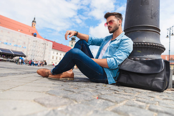 homem casual vestindo óculos de sol vermelhos e camisa azul sentado perto do poste de iluminação no fundo urbano olhando para o lado, imagem de comprimento total
 - Foto, Imagem