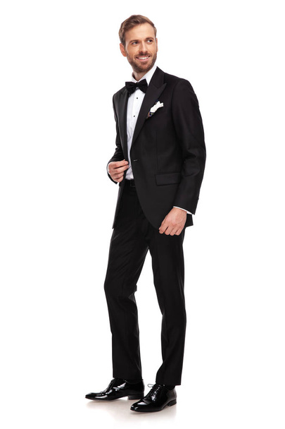 handsome businessman in black suit looks to side while standing on white background and holding collar, full body picture - Fotoğraf, Görsel