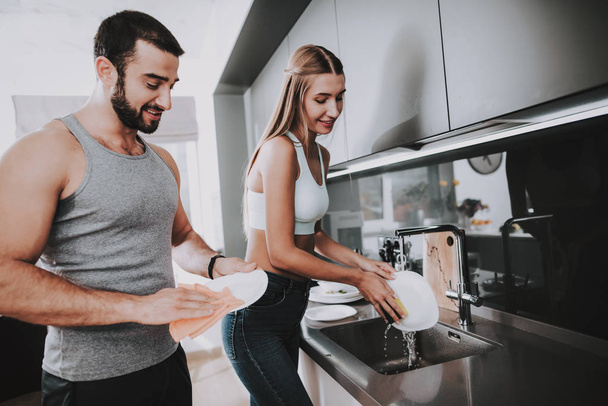 Sporty Couple Is Washing A Dish At The Kitchen. Dishware Cleaning. Working Together. Domestic Life. Tableware In The Sink. Shiny Kitchen. Holiday Leisure. Weekend Activity. Sunny Day. - Foto, immagini