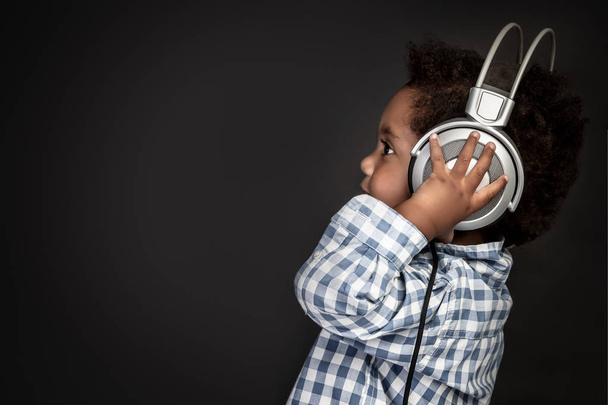 Profile portrait of a little curly African-American baby boy wearing big headphones and listening favourite music over black background, sweet child having fun - Foto, Bild